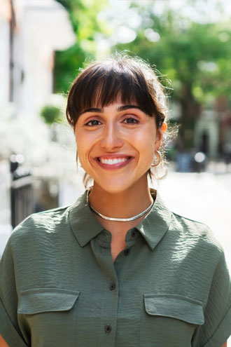 A woman with medium brown hair and bangs smiles outdoors, wearing a green button-up shirt and a silver necklace. The background is blurred with greenery and buildings visible.