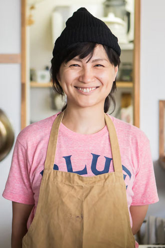 A smiling person wearing a black beanie, pink T-shirt, and tan apron stands indoors, possibly in a kitchen setting.